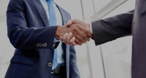 Close-up photo of two people in dark business suits shaking hands, symbolizing cooperation or agreement.